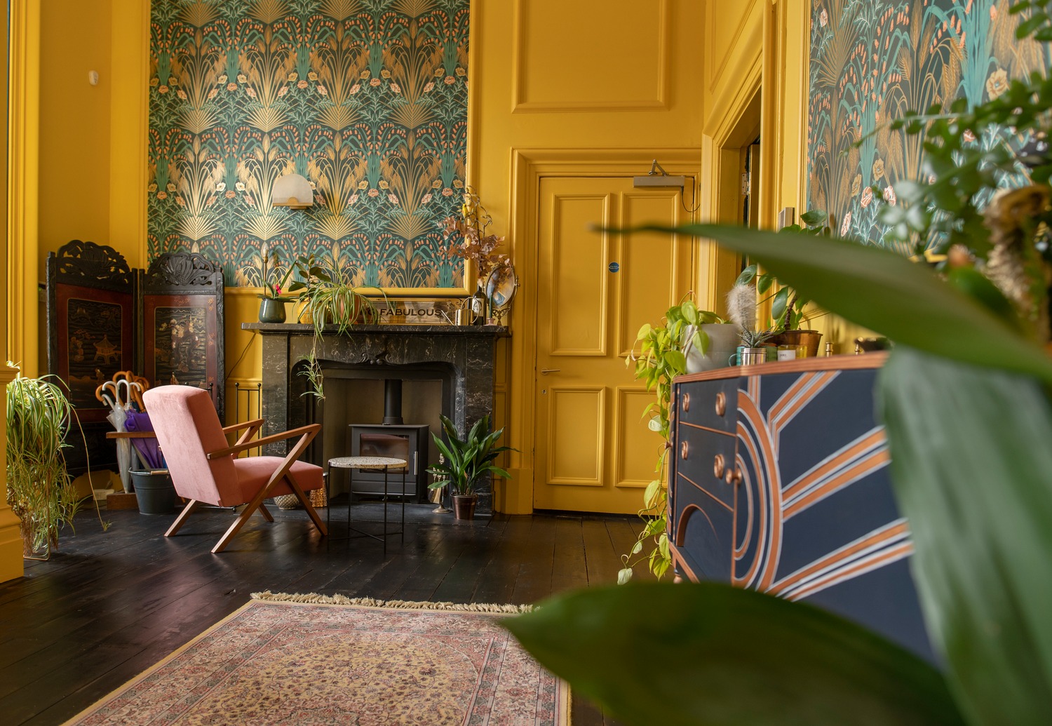 The bright yellow entrance hall at Netherbyres, with blue art deco wallpaper and sideboard and a marble fireplace.